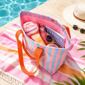 May include: A tote bag with vertical light blue and pink stripes and orange handles. The interior contains a rolled pink and white striped towel, a Sunbound sunscreen tube, a pink water bottle, sunglasses, and a book titled Coastal Summer Escapes.