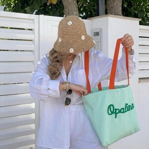 May include: A tan crocheted bucket hat with small white flower appliqu&eacute;s and a light green tote bag with orange straps and the word Opaline embroidered in dark green script. The bag features a fine vertical ribbed texture across the surface.