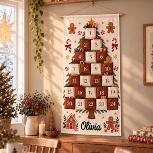 Può includere: Un calendario dell'Avvento da parete in tessuto bianco con un disegno di albero di Natale stampato e ventiquattro tasche numerate che si alternano tra i colori marrone e crema. Il nome Olivia è stampato in basso con grafiche di omini di pan di zenzero.