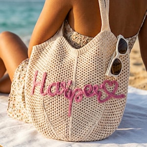 May include: A cream-colored, open-weave tote bag with the name "Harper" embroidered in pink script. White sunglasses are tucked into the bag's strap. The bag is on a beach.