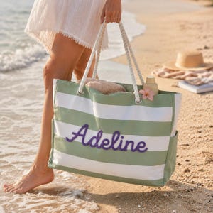 May include: A rectangular tote bag featuring horizontal sage green and white stripes with the name Adeline embroidered in purple script on the front. The bag includes thick white rope handles attached through silver metal grommets.