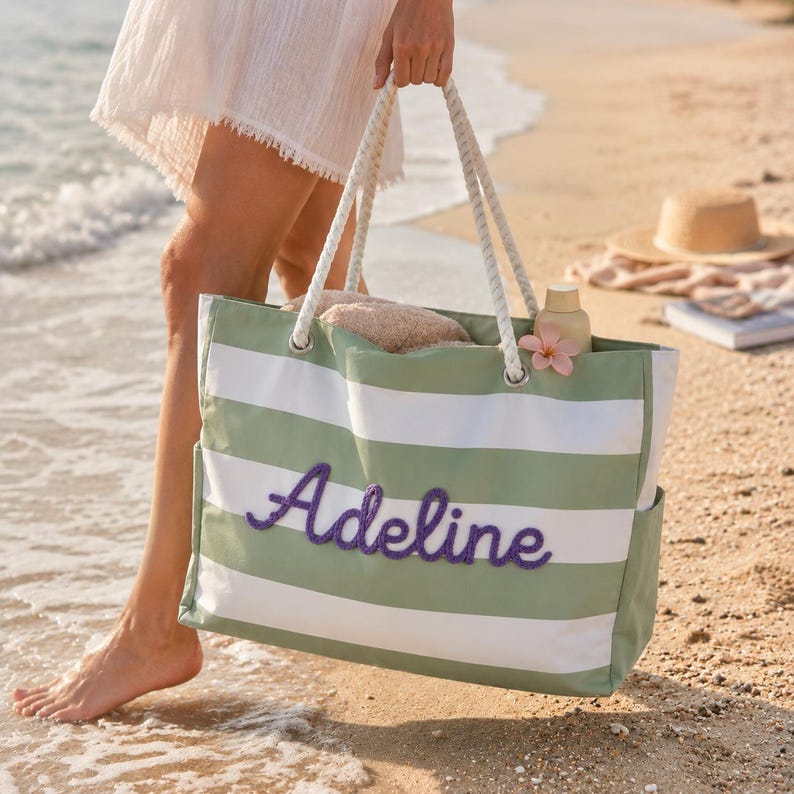 May include: A rectangular tote bag featuring horizontal sage green and white stripes with the name Adeline embroidered in purple script on the front. The bag includes thick white rope handles attached through silver metal grommets.