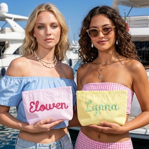 Puede incluir: Dos mujeres sostienen neceseres personalizados. Una bolsa es rosa con el nombre "Elowen" bordado en rojo, la otra es amarilla con "Lynna" en verde. Ambas bolsas tienen un patr&oacute;n de rayas.