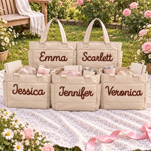 Peut inclure: Cinq sacs fourre-tout matelassés beiges avec poches latérales, chacun portant un nom brodé en marron : Emma, Scarlett, Jessica, Jennifer et Veronica. Les sacs ont deux anses et sont disposés sur une surface en dentelle blanche en extérieur.