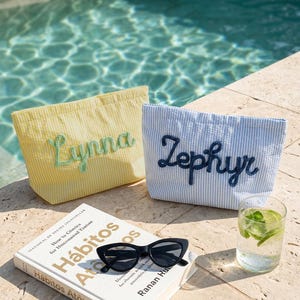Puede incluir: Dos bolsas de mano de seersucker, una amarilla con "Lynna" bordado en verde, la otra a rayas azules y blancas con "Zephyr" en azul marino. Un libro, gafas de sol negras y un vaso de agua con lima y menta tambi&eacute;n est&aacute;n presentes.