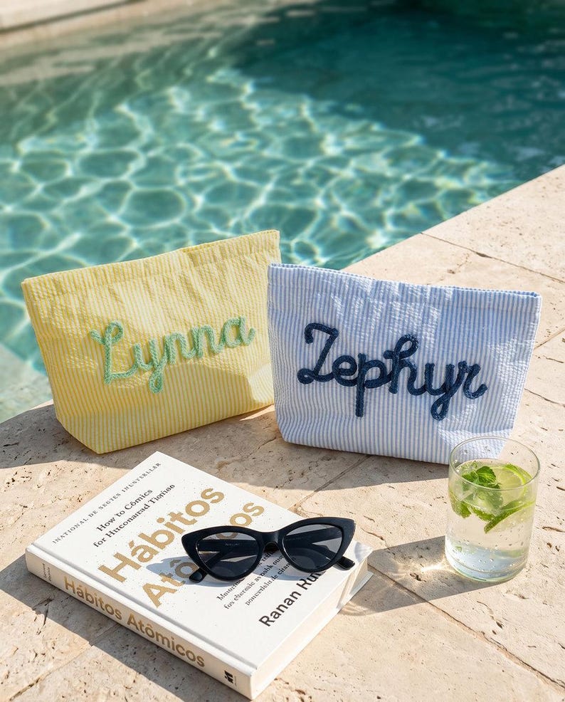 Puede incluir: Dos bolsas de mano de seersucker, una amarilla con "Lynna" bordado en verde, la otra a rayas azules y blancas con "Zephyr" en azul marino. Un libro, gafas de sol negras y un vaso de agua con lima y menta tambi&eacute;n est&aacute;n presentes.