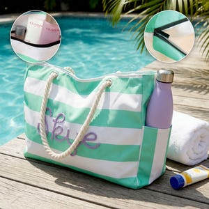 May include: A mint green and white striped canvas tote bag with thick white rope handles and the name Skye embroidered in purple script on the front. The bag features a side pocket holding a lavender water bottle, a zipper closure, and an interior.