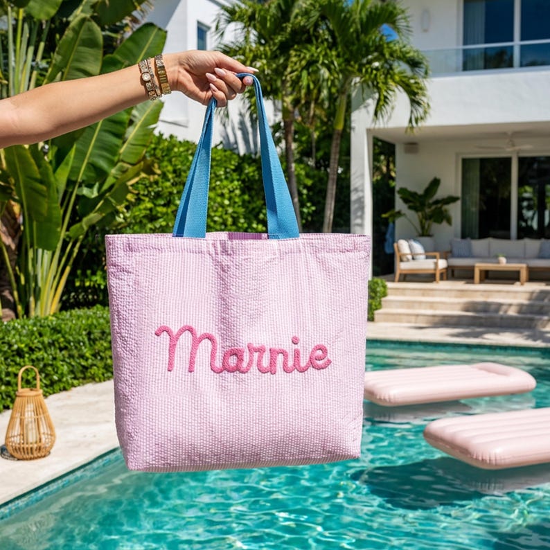 May include: A light pink seersucker tote bag featuring two blue fabric handles and the name Marnie embroidered in a cursive pink font on the front. The bag has a rectangular shape and a textured fabric surface, providing a simple and functional design.