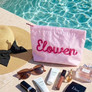 Puede incluir: Un conjunto de productos de belleza y cuidado solar, que incluye un neceser rosa a rayas con el nombre "Elowen" bordado, gafas de sol, protector solar, l&aacute;piz labial, brillo labial, perfume y un sombrero para el sol.
