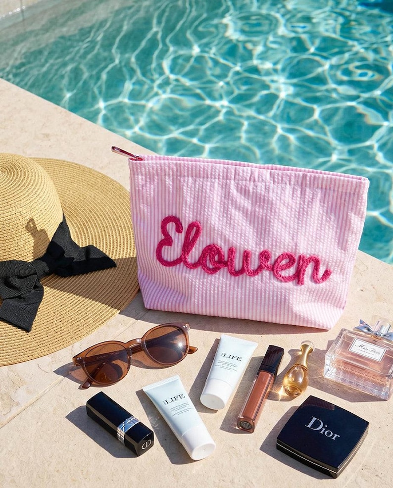 Puede incluir: Un conjunto de productos de belleza y cuidado solar, que incluye un neceser rosa a rayas con el nombre "Elowen" bordado, gafas de sol, protector solar, l&aacute;piz labial, brillo labial, perfume y un sombrero para el sol.