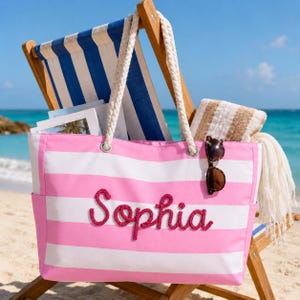 May include: A pink and white striped canvas tote bag featuring the name Sophia in raised pink rope lettering. The bag has thick white rope handles and a pair of dark brown sunglasses hanging from one of the handles, set against a beach backdrop.