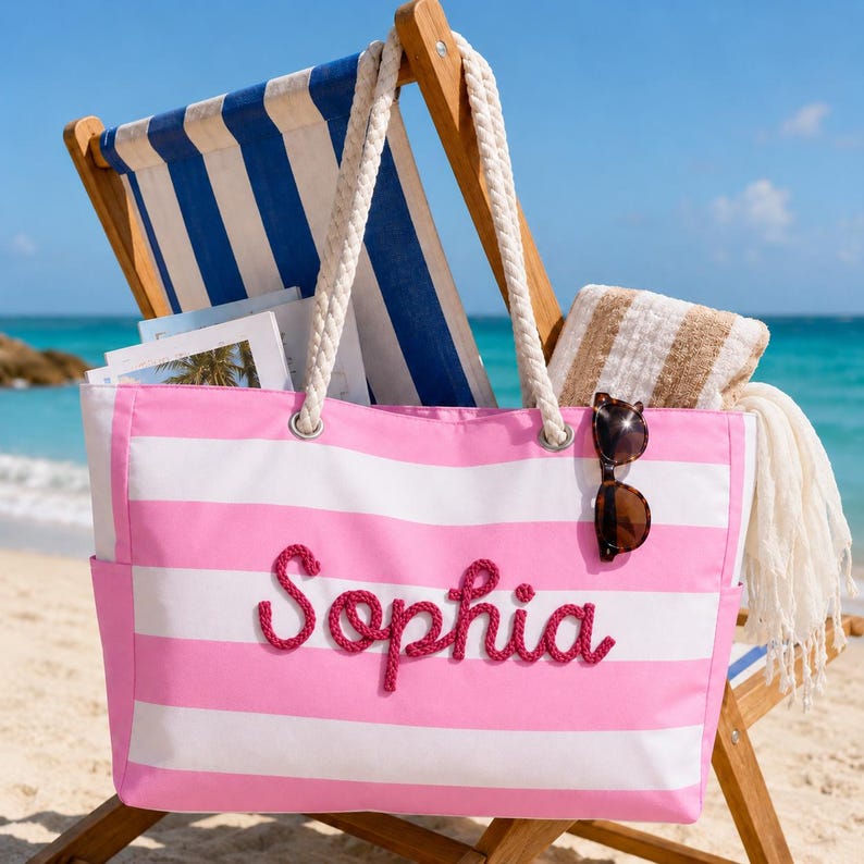 May include: A pink and white striped canvas tote bag featuring the name Sophia in raised pink rope lettering. The bag has thick white rope handles and a pair of dark brown sunglasses hanging from one of the handles, set against a beach backdrop.