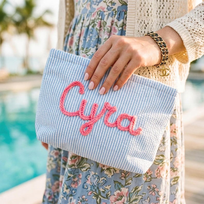 Puede incluir: Un bolso de mano a rayas azules y blancas con el nombre "Cyra" bordado en rosa en cursiva. El bolso de mano lo sostiene una persona que lleva un vestido floral y una rebeca color crema. Se ve una pulsera de cadena dorada y negra.