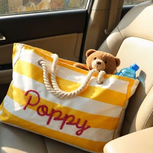 May include: A yellow and white striped canvas tote bag with thick rope handles and the name Poppy embroidered in red script on the front. Inside the bag sits a brown plush teddy bear and a clear plastic water bottle with a blue cap.