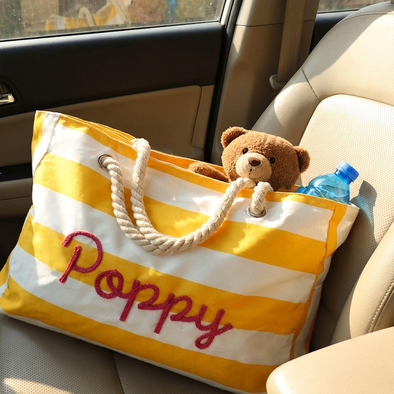 May include: A yellow and white striped canvas tote bag with thick rope handles and the name Poppy embroidered in red script on the front. Inside the bag sits a brown plush teddy bear and a clear plastic water bottle with a blue cap.
