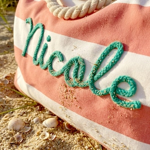 May include: A canvas tote bag featuring horizontal coral pink and white stripes with a thick, cream-colored rope handle. The name Nicole is embroidered across the front in a cursive script using a textured, bright teal rope-like material.