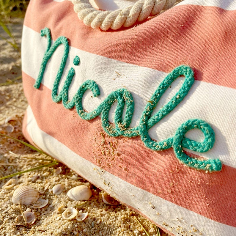 May include: A canvas tote bag featuring horizontal coral pink and white stripes with a thick, cream-colored rope handle. The name Nicole is embroidered across the front in a cursive script using a textured, bright teal rope-like material.