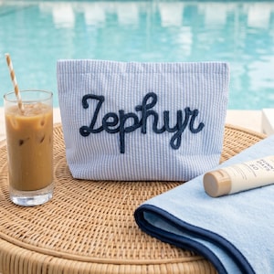 Puede incluir: Un bolso tote a rayas azules y blancas con el nombre "Zephyr" bordado en azul marino. La bolsa est&aacute; sobre una mesa de mimbre junto a un vaso de caf&eacute; helado y un tubo de protector solar.