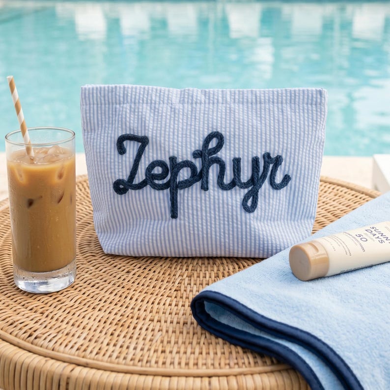Puede incluir: Un bolso tote a rayas azules y blancas con el nombre "Zephyr" bordado en azul marino. La bolsa est&aacute; sobre una mesa de mimbre junto a un vaso de caf&eacute; helado y un tubo de protector solar.