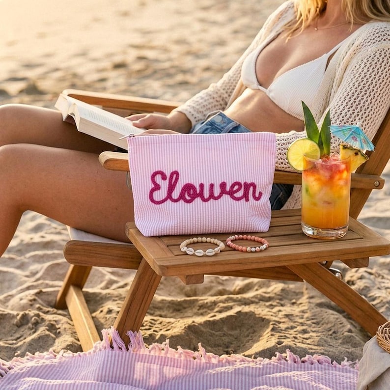 Puede incluir: Un neceser a rayas rosas y blancas con el nombre "Elowen" bordado en cursiva. La bolsa est&aacute; sobre una mesa de madera junto a un c&oacute;ctel y dos pulseras. Escena de playa con una persona al fondo.