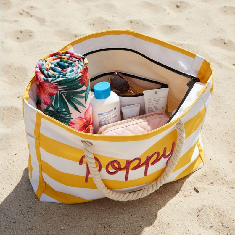May include: A yellow and white striped canvas tote bag with rope handles and the name Poppy embroidered in red script on the front. The bag is filled with a rolled floral towel, a bottle of sunscreen, sunglasses, and a small quilted pouch.