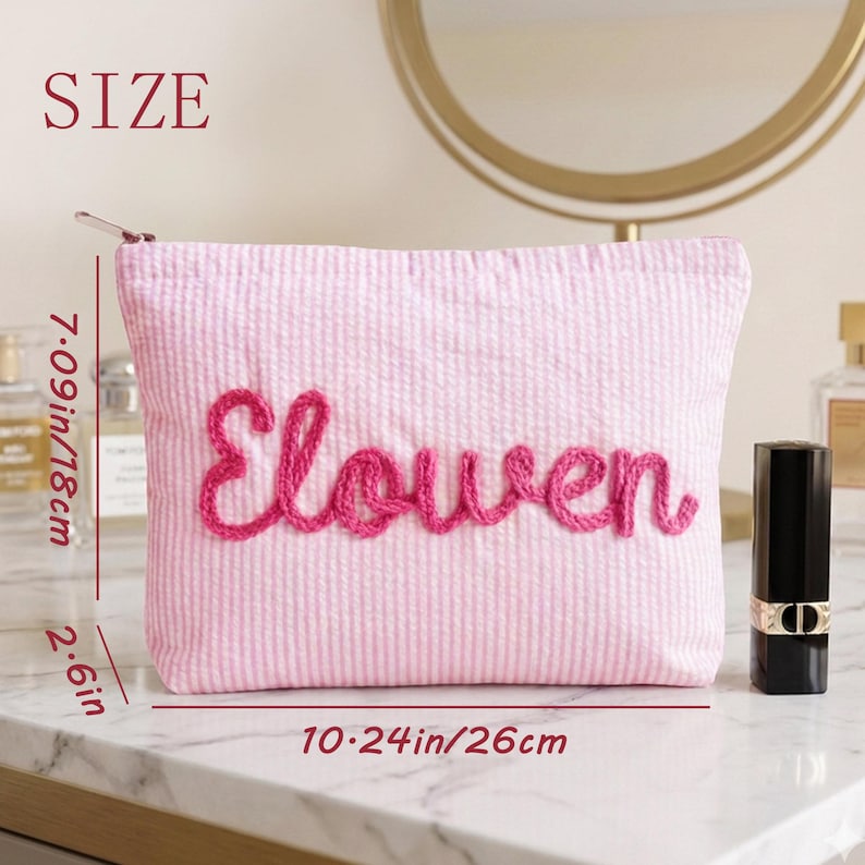 Puede incluir: Bolsa de cosm&eacute;ticos rosa de seersucker con el nombre "Elouen" bordado en cursiva. La bolsa mide 18 cm de alto, 26 cm de ancho y 6,6 cm de profundidad. Un tubo de l&aacute;piz labial negro est&aacute; al lado de la bolsa.