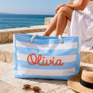 May include: A large rectangular tote bag featuring horizontal light blue and white stripes. The bag has thick white rope handles attached with silver metal grommets and the name Olivia embroidered in bright orange rope on the front panel.