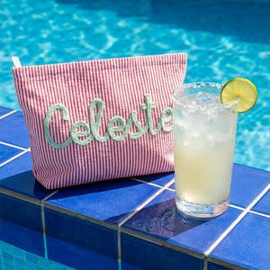 Puede incluir: Un neceser a rayas rojas y blancas con el nombre "Celeste" bordado en verde. Una margarita con una rodaja de lima est&aacute; junto a la bolsa sobre baldosas azules, con una piscina al fondo.
