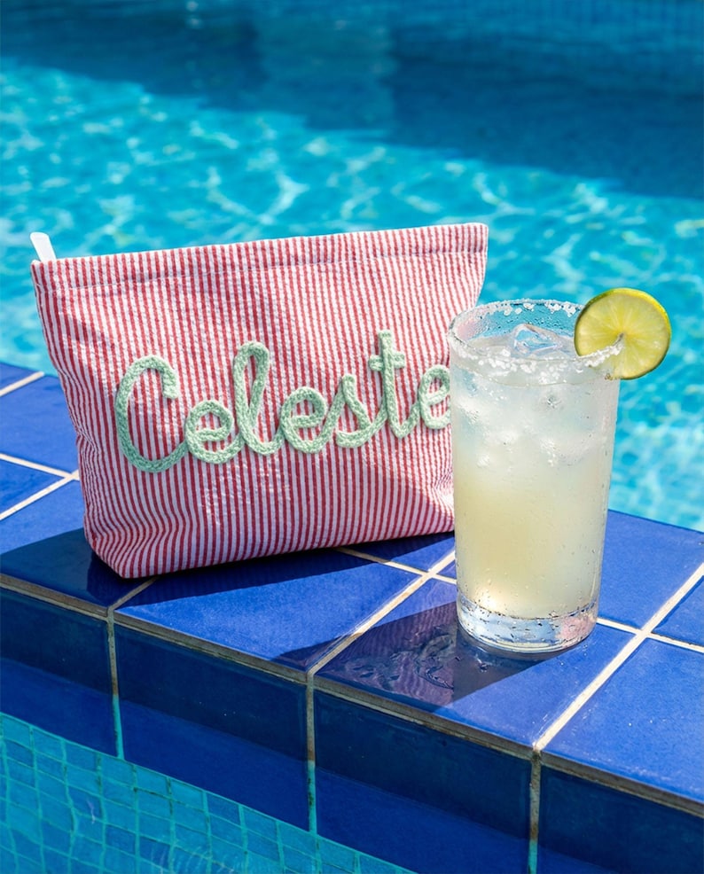 Puede incluir: Un neceser a rayas rojas y blancas con el nombre "Celeste" bordado en verde. Una margarita con una rodaja de lima est&aacute; junto a la bolsa sobre baldosas azules, con una piscina al fondo.