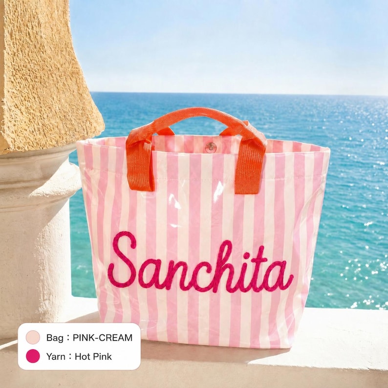 K&ouml;nnte beinhalten: Eine Tragetasche mit vertikalen rosa und cremefarbenen Streifen und zwei orangefarbenen Griffen. Der Name Sanchita ist in kr&auml;ftigem Pink in Schreibschrift auf die Vorderseite gestickt. Eine kleine Farblegende zeigt, dass die Tasche rosa-creme ist.