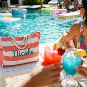 May include: A rectangular tote bag featuring horizontal coral pink and white stripes with thick white rope handles. The name Nicole is embroidered in a dark teal cursive script on the front of the bag, which sits on a white surface.