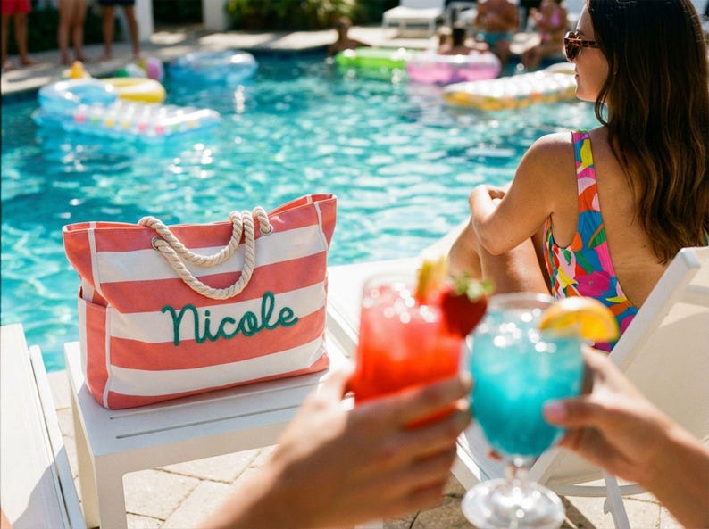 May include: A rectangular tote bag featuring horizontal coral pink and white stripes with thick white rope handles. The name Nicole is embroidered in a dark teal cursive script on the front of the bag, which sits on a white surface.