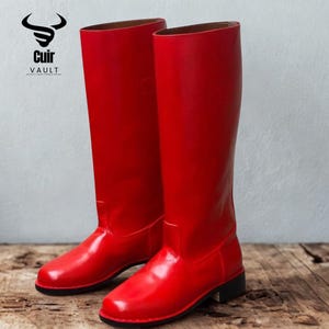 Puede incluir: Un par de botas rojas altas y brillantes con un acabado liso. Las botas tienen un diseño clásico, con punta redonda y tacón bajo negro. Las botas se exhiben sobre una superficie de madera. El logotipo de la marca "Cuir Vault" es visible.
