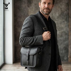 Puede incluir: Bolso mensajero de cuero negro con cierre de solapa y correa ajustable para el hombro. El bolso tiene una hebilla y un pequeño acento rojo. El logotipo "Cuir Vault" es visible en la esquina superior izquierda.