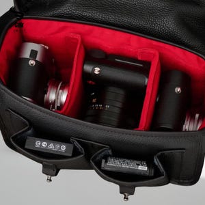 May include: Black leather camera bag with a red interior. The bag is open, revealing multiple compartments holding cameras and lenses. Two small pockets on the front of the bag hold batteries. The bag has the words "Cuir Vault" printed on the top left corner.