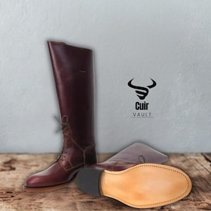 May include: A pair of tall, burgundy leather riding boots. One boot is upright, laced up, and the other is lying down, showcasing the sole. The boots have a classic design, with a smooth finish and a subtle sheen. The brand logo "Cuir Vault" is visible.