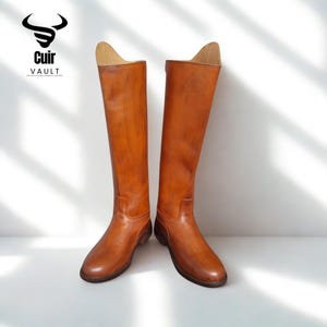 May include: A pair of brown leather riding boots, knee-high with a polished finish. The boots have a rounded toe and are displayed against a white background. The logo "Cuir Vault" is visible in the upper left corner. The boots are a classic equestrian style.