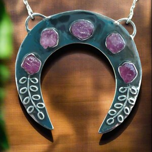 May include: A silver crescent moon pendant necklace with five pink gemstones set into the metal. The pendant has a floral design on each side.