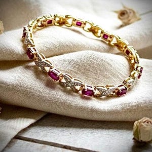 May include: A gold-toned bracelet with alternating ruby-colored rectangular stones and diamond-like accents. The bracelet is displayed on a cream-colored fabric, with dried rose buds in the background.