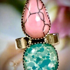 May include: A silver ring featuring two stones. The top stone is a pink, teardrop-shaped stone with dark veining. The bottom stone is a square, turquoise-colored stone. Both stones are set in a silver bezel.