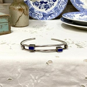 May include: A silver-toned cuff bracelet with two square, cobalt blue accents. The bracelet is displayed on a white, patterned surface, with vintage blue and white china in the background. The bracelet is a simple, yet elegant piece of jewelry.