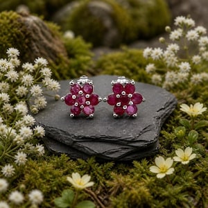 May include: A pair of stud earrings featuring a cluster of seven round-cut, deep pinkish-red rubies arranged in a floral pattern. Each ruby is set in a polished silver-toned metal prong setting, creating a symmetrical flower shape for each earring.