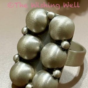 May include: A silver-toned ring featuring a rectangular face adorned with six large, smooth, spherical domes arranged in two columns, accented by smaller spherical beads between them. The text The Wishing Well is visible above the ring.