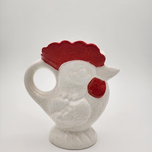 May include: A white ceramic pitcher shaped like a rooster. The pitcher has a red comb and wattle, with a speckled finish. The handle is a loop, and the base is a small pedestal.