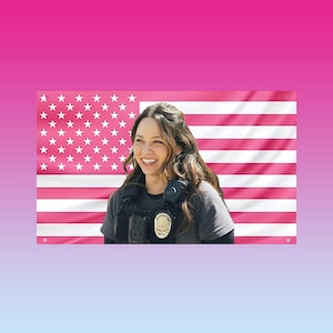 May include: A pink American flag with white stars and stripes is in the background. A person wearing a gray shirt and a badge is smiling in front of the flag. The background is a gradient of pink and blue.