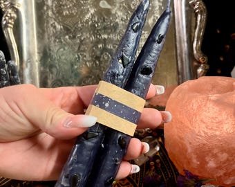 May include: Two dark blue taper candles with black accents are held together with a decorative band. The candles are displayed against a backdrop of a silver tray, tarot cards, and dried flowers. The candles are approximately 8 inches in length.