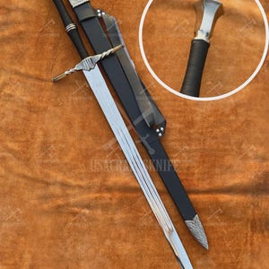 May include: A long, silver sword with a black handle and a matching black sheath. The sword has a detailed crossguard and a decorative pommel. The sheath is made of leather and has silver accents. The text "USACRA KNIFE" is visible.
