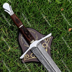 May include: A detailed image of a decorative sword with a patterned blade and a wooden handle. The handle features intricate carvings and is accented with silver-colored metal. The sword rests on a brown stand with gold-colored accents and text. The background is green grass.