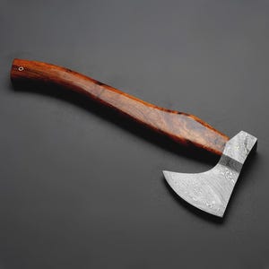 May include: A handcrafted axe with a dark wooden handle and a patterned steel blade. The axe head has a curved blade and a textured surface. The handle is a rich brown color, and the axe is set against a dark background.