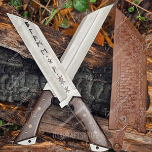 May include: Two crossed, large, silver blades with dark wooden handles and decorative rivets. The blades have etched runes and symbols. A brown leather sheath with a woven pattern is visible. The knives are displayed on a bed of wood and leaves.